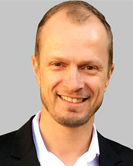 Headshot of a person with short hair wearing a dark blazer, light background