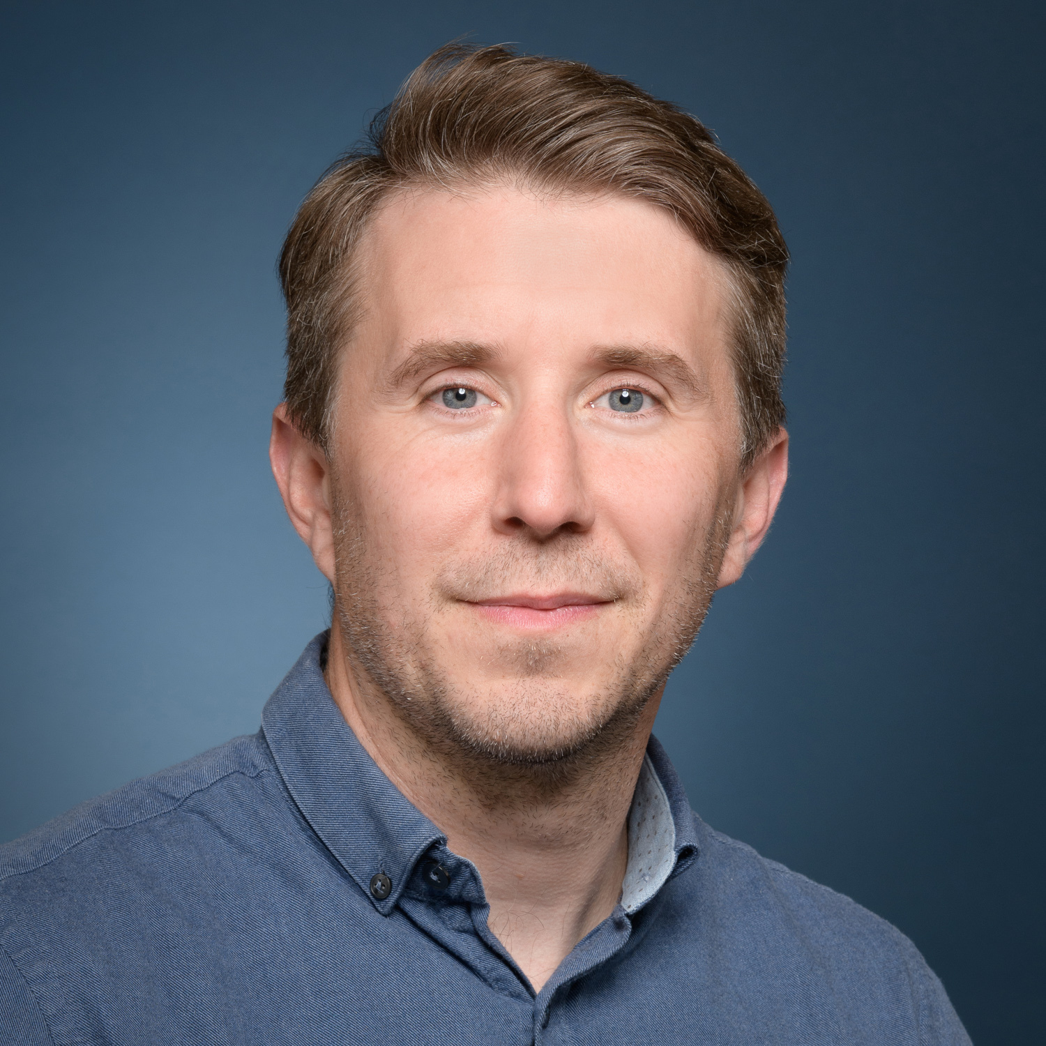 Headshot of a person with short brown hair and light eyes, wearing a blue button-up shirt